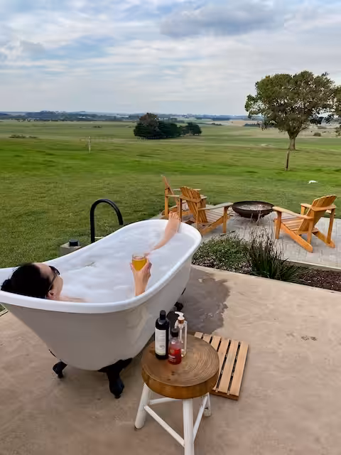 A woman reclines in an outdoor bath tub with a beer, overlooking paddocks.